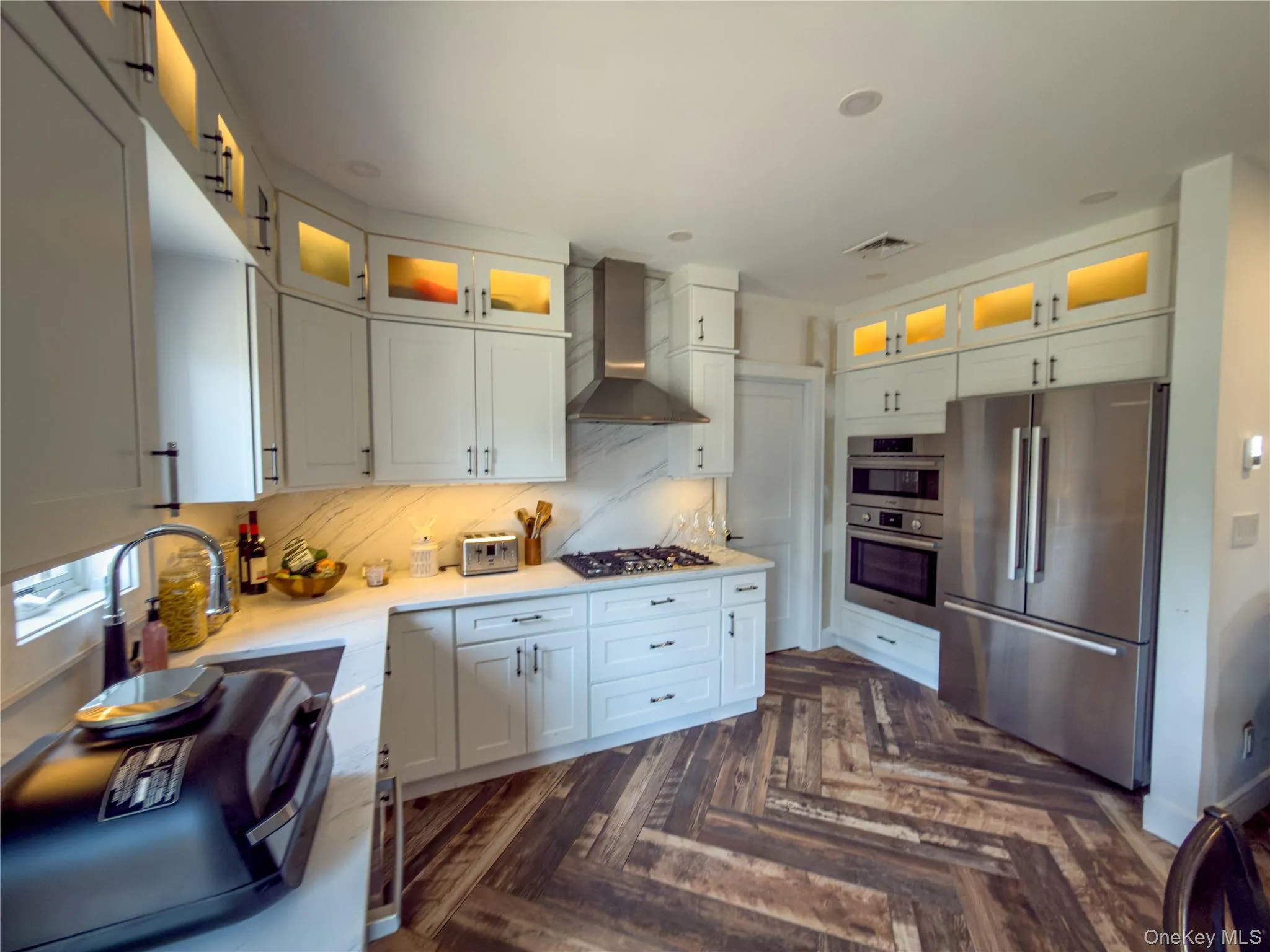 White cabinetry with integrated lighting, stainless steel appliances, herringbone flooring, and cooktop with a stainless steel range hood White cabinetry with integrated lighting, stainless steel appliances, herringbone flooring, and cooktop with a stainless steel range hood