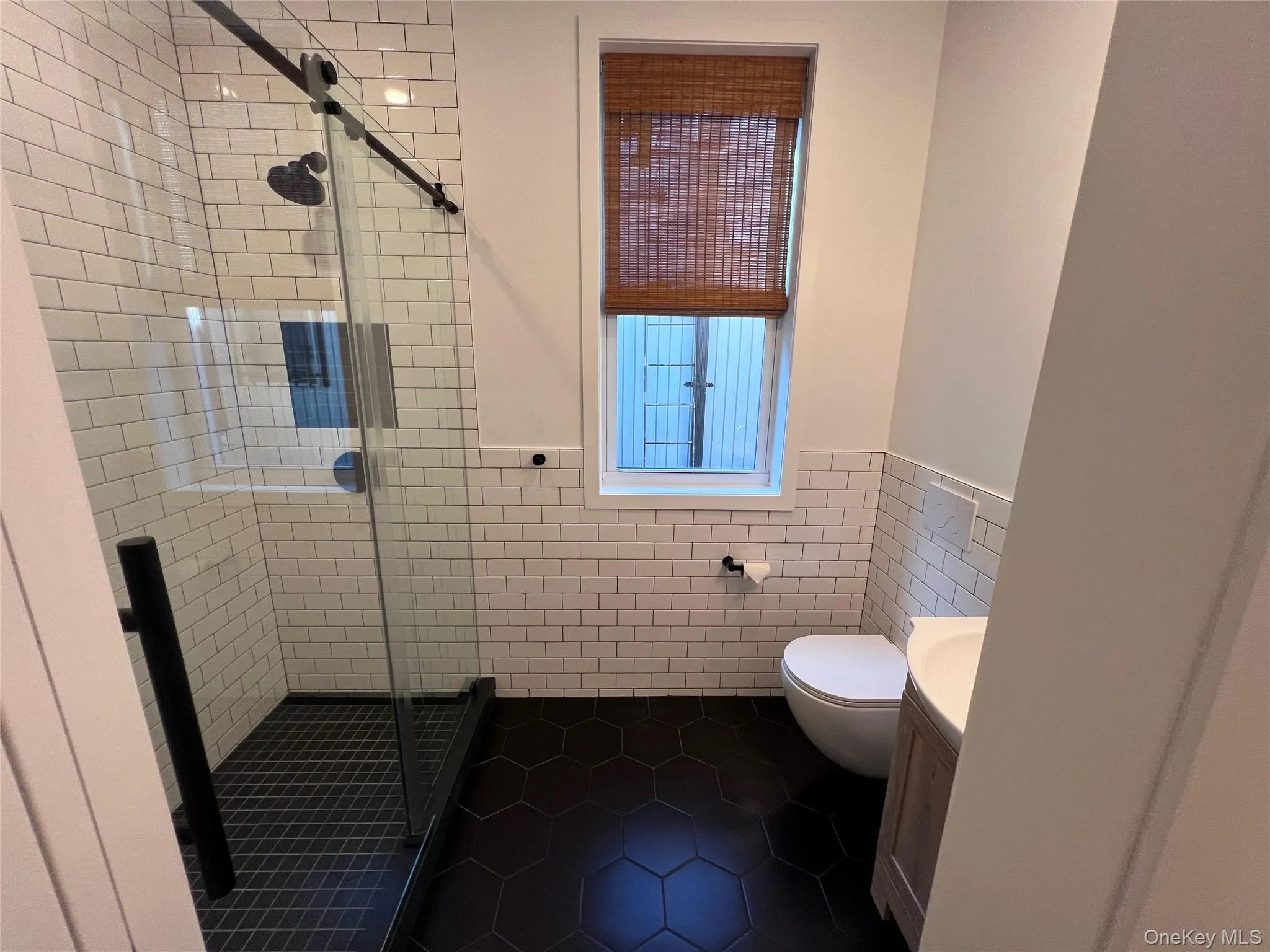 Glass-enclosed shower with black hardware, white subway tile surround, dark hexagonal floor tiles, and a floating vanity Glass-enclosed shower with black hardware, white subway tile surround, dark hexagonal floor tiles, and a floating vanity
