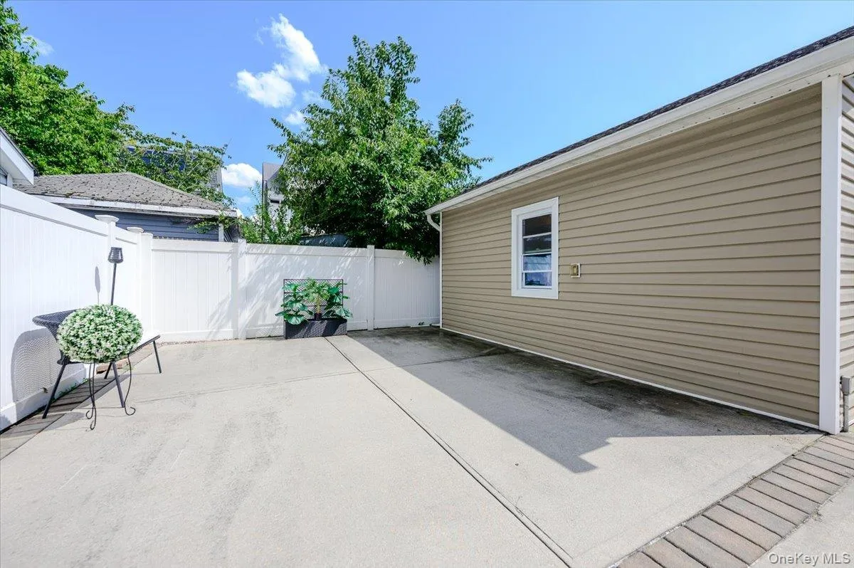Expansive concrete patio with mature landscaping and white privacy fencing, adjoining a single-story residence with tan fiber cement siding and a contemporary window design Expansive concrete patio with mature landscaping and white privacy fencing, adjoining a single-story residence with tan fiber cement siding and a contemporary window design
