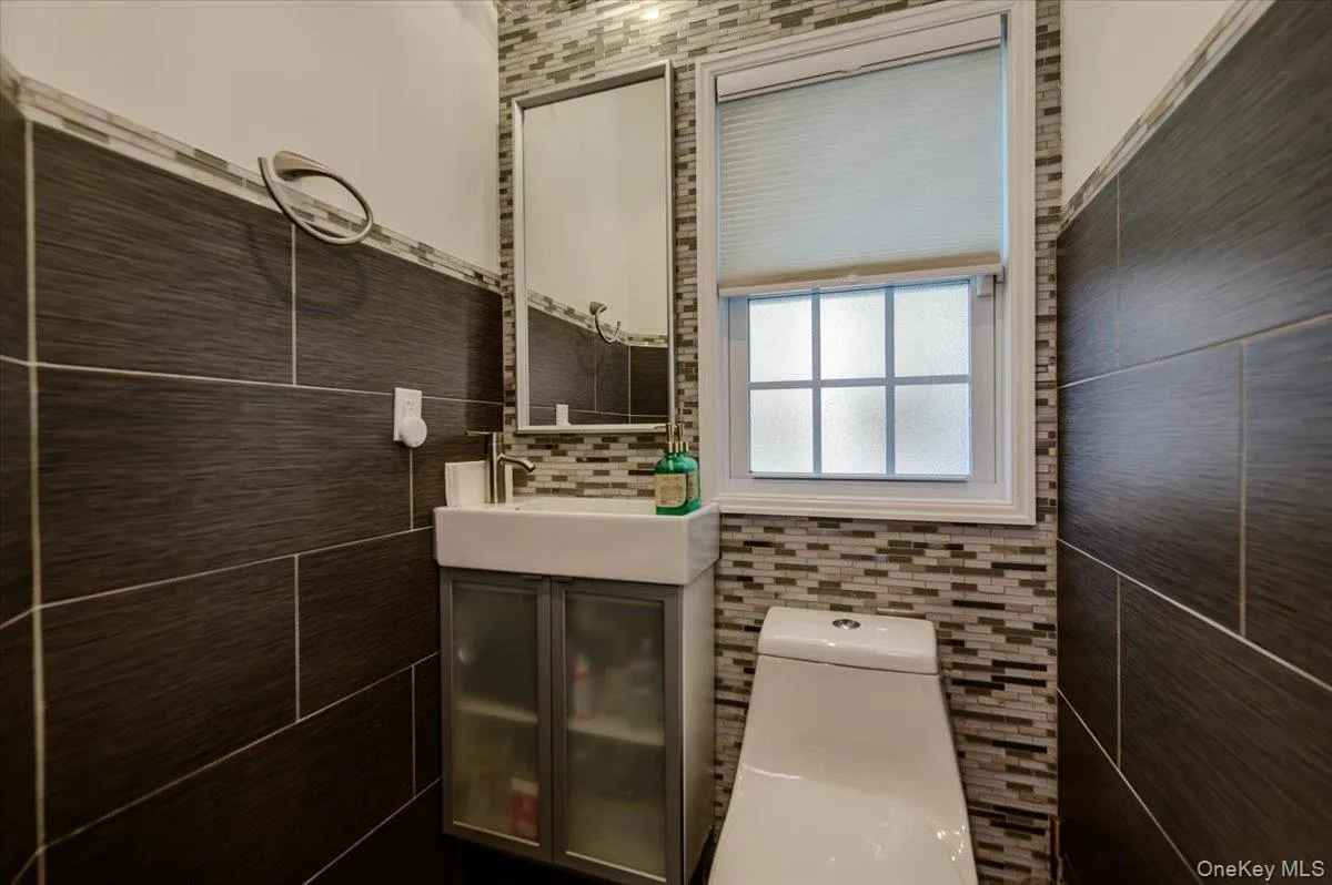 Tiled accent wall with mosaic detailing, rectangular window with privacy glass, wall-mounted vanity with integrated sink, and modern toilet fixture Tiled accent wall with mosaic detailing, rectangular window with privacy glass, wall-mounted vanity with integrated sink, and modern toilet fixture