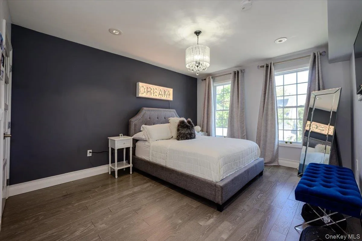 Spacious primary bedroom featuring a dramatic navy accent wall, wood-finish flooring throughout, and abundant natural light from multiple windows dressed in neutral curtains Spacious primary bedroom featuring a dramatic navy accent wall, wood-finish flooring throughout, and abundant natural light from multiple windows dressed in neutral curtains