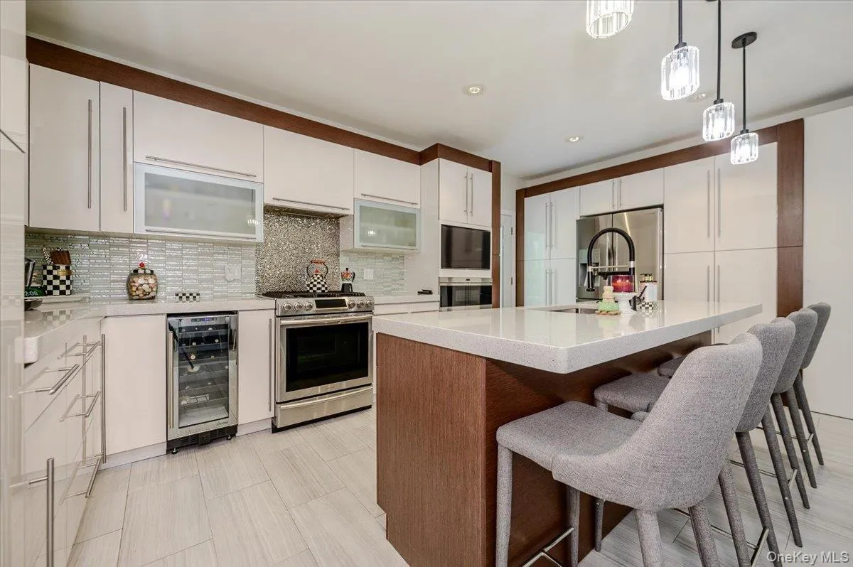 White flat-panel cabinetry with wood-finish accents, stainless steel appliances, a center island with countertop seating, a beverage refrigerator, and a mosaic tile backsplash White flat-panel cabinetry with wood-finish accents, stainless steel appliances, a center island with countertop seating, a beverage refrigerator, and a mosaic tile backsplash