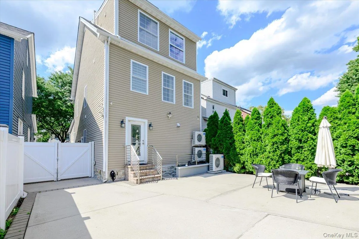 Expansive concrete patio with mature evergreen privacy screening, complemented by a contemporary two-story colonial residence featuring horizontal siding, multi-pane windows, and a covered entry Expansive concrete patio with mature evergreen privacy screening, complemented by a contemporary two-story colonial residence featuring horizontal siding, multi-pane windows, and a covered entry