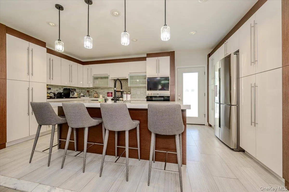 Kitchen featuring an expansive island with countertop seating, modern pendant lighting, integrated stainless-steel refrigerator, ample white cabinetry, and wood-finish accents Kitchen featuring an expansive island with countertop seating, modern pendant lighting, integrated stainless-steel refrigerator, ample white cabinetry, and wood-finish accents