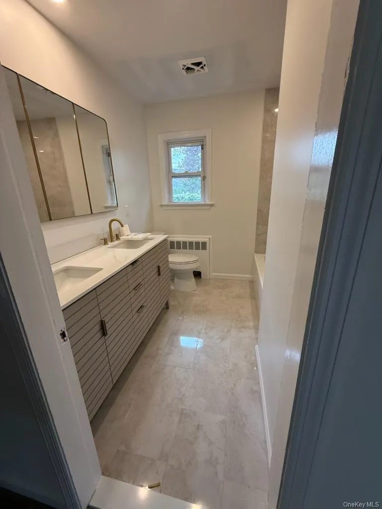 Bathroom with radiator, double vanity, marble look tiles, toilet, and baseboards Bathroom with radiator, double vanity, marble look tiles, toilet, and baseboards
