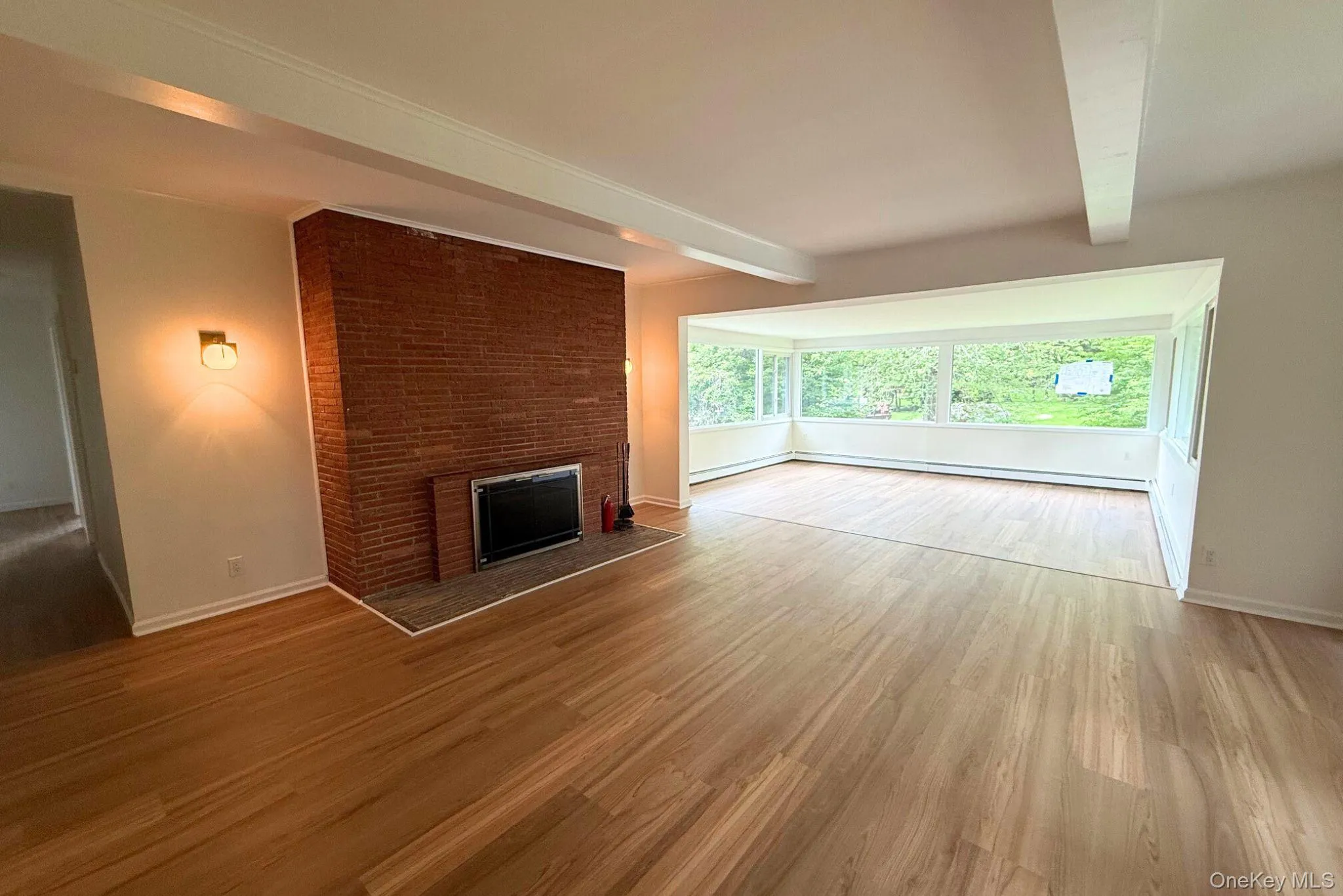 Unfurnished living room with wood finished floors, a brick fireplace, baseboards, beam ceiling, and a baseboard heating unit Unfurnished living room with wood finished floors, a brick fireplace, baseboards, beam ceiling, and a baseboard heating unit