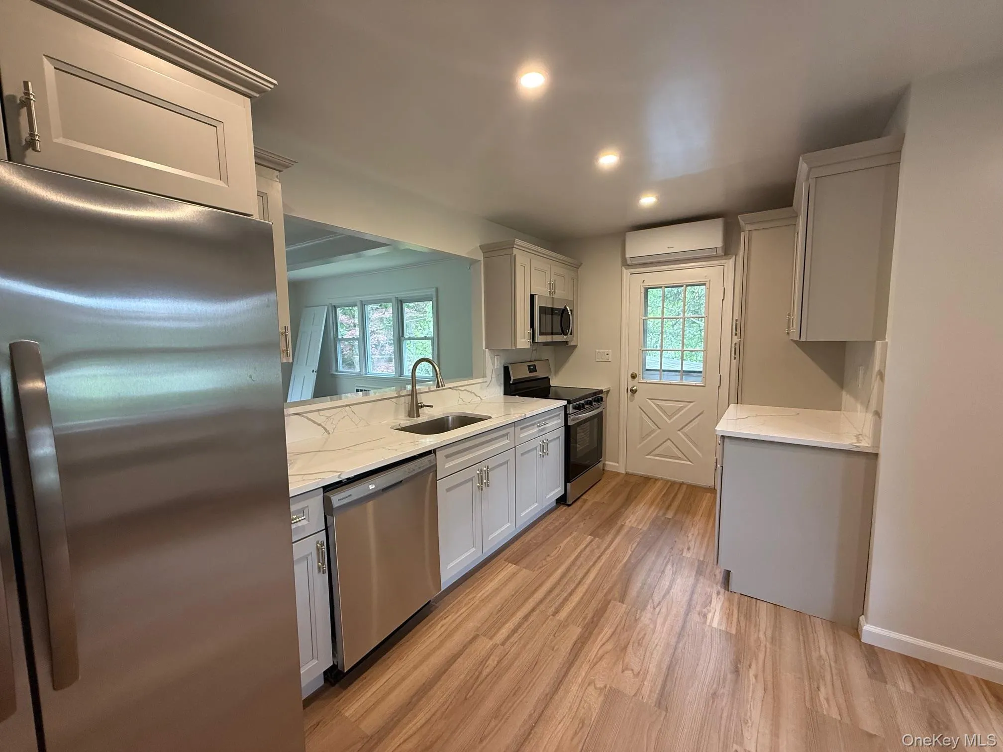 Kitchen featuring appliances with stainless steel finishes, a wall mounted air conditioner, a sink, light wood-type flooring, and light stone countertops Kitchen featuring appliances with stainless steel finishes, a wall mounted air conditioner, a sink, light wood-type flooring, and light stone countertops