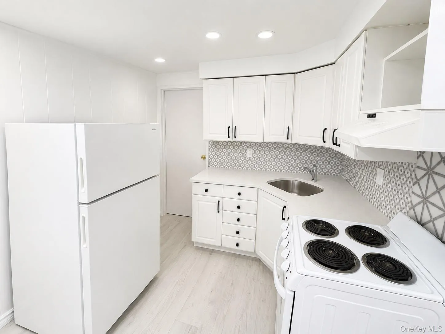 White cabinetry with matte black hardware, geometric pattern tile backsplash, light-toned wood-finish flooring, recessed ceiling lighting, and white appliances White cabinetry with matte black hardware, geometric pattern tile backsplash, light-toned wood-finish flooring, recessed ceiling lighting, and white appliances