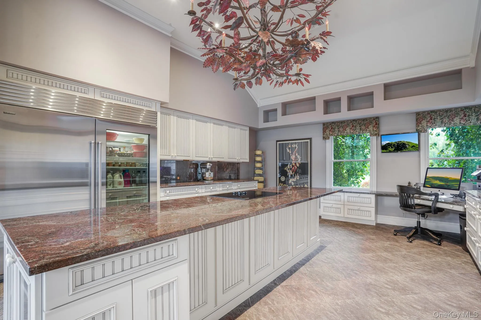 Vaulted ceiling with an ornate chandelier, expansive stone island, integrated stainless steel refrigerator, white cabinetry, and built-in desk area. Vaulted ceiling with an ornate chandelier, expansive stone island, integrated stainless steel refrigerator, white cabinetry, and built-in desk area.