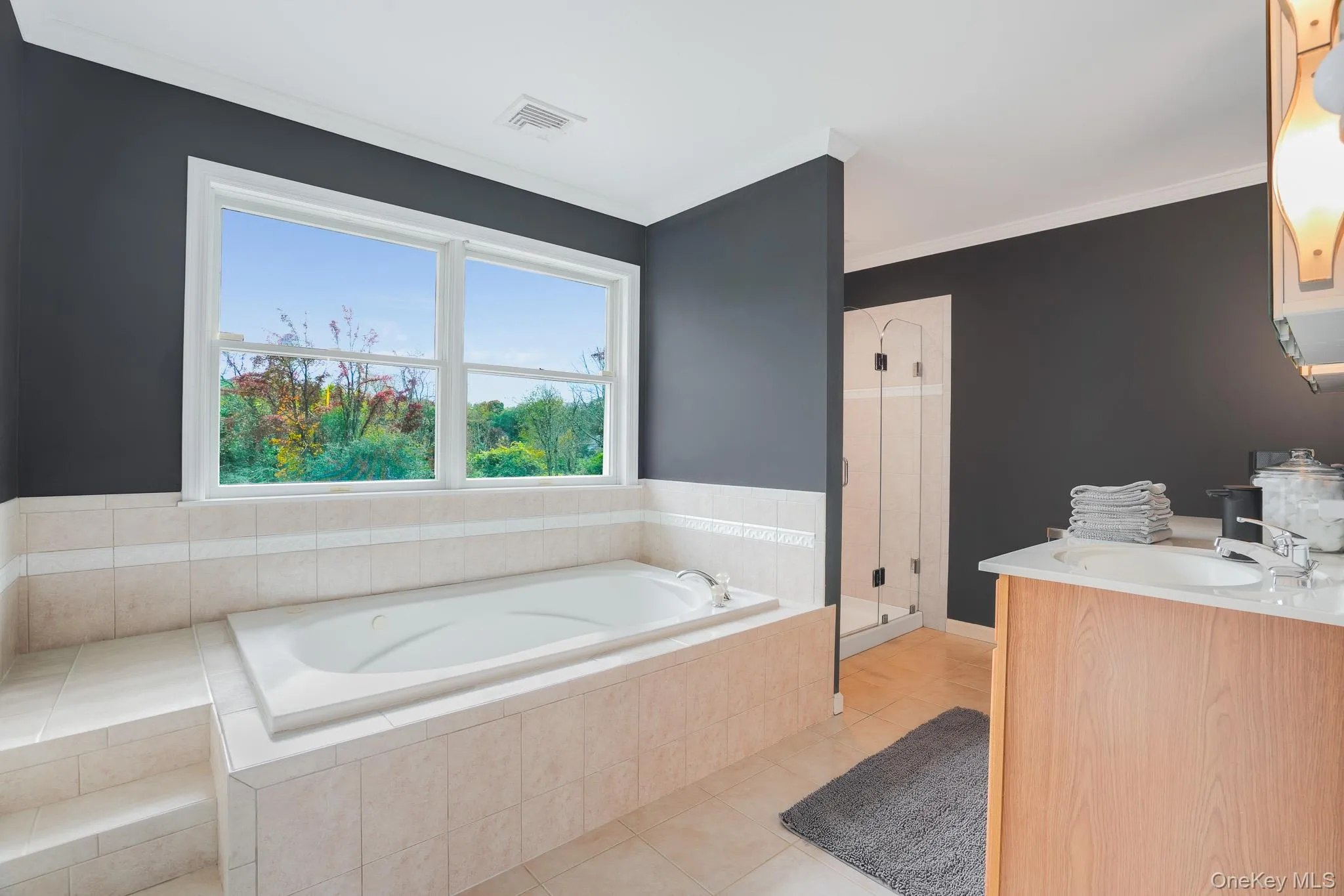 Deep soaking tub with tiled surround, walk-in shower with glass enclosure, wood-finish vanity with countertop sink, and a large window Deep soaking tub with tiled surround, walk-in shower with glass enclosure, wood-finish vanity with countertop sink, and a large window