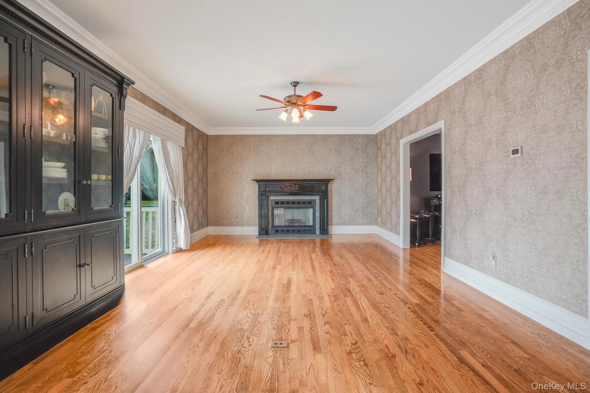 Hardwood flooring, fireplace with dark mantel, patterned wall covering, ceiling fan, and built-in cabinetry with glass doors. Hardwood flooring, fireplace with dark mantel, patterned wall covering, ceiling fan, and built-in cabinetry with glass doors.