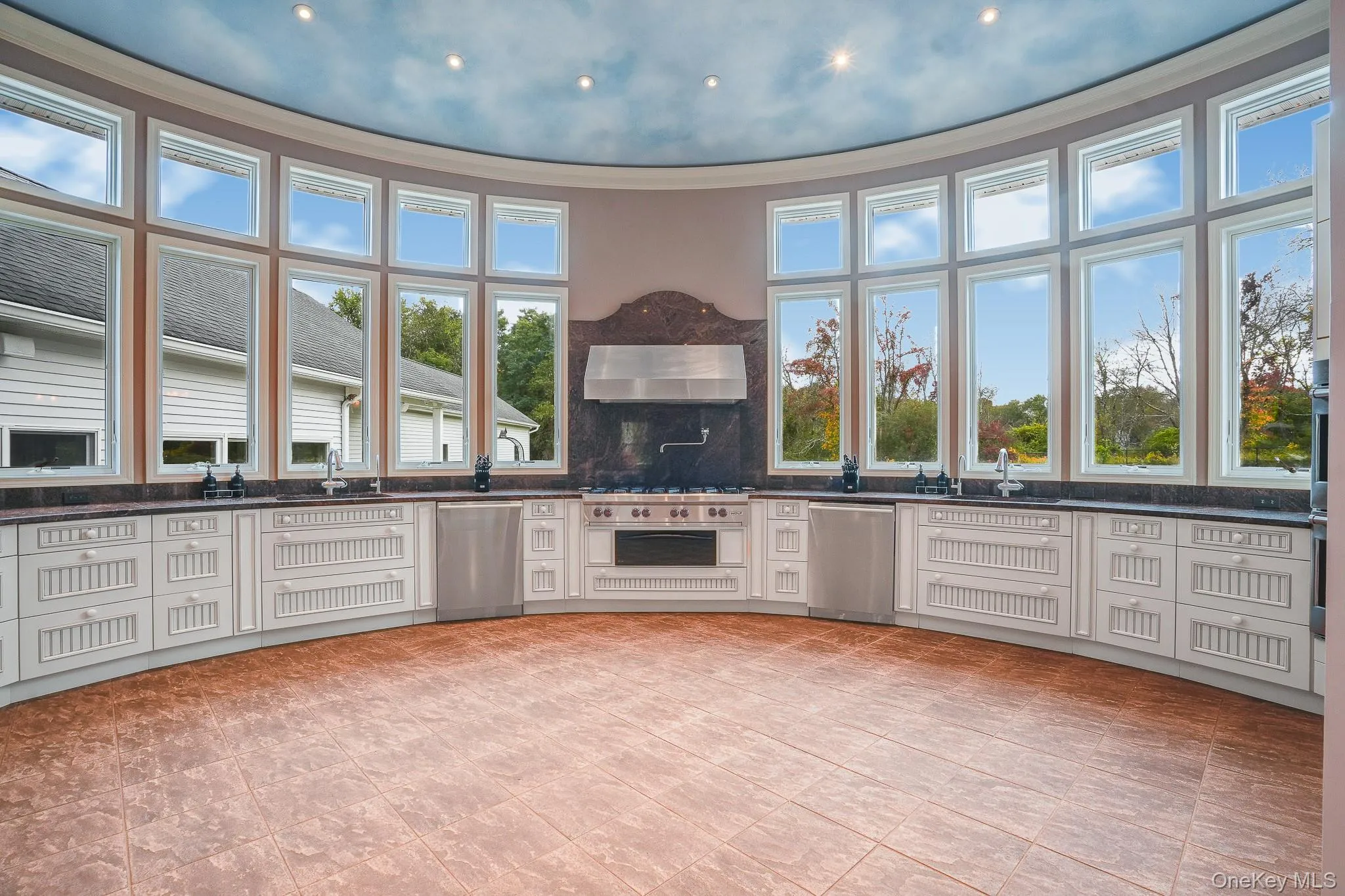 Curved wall of expansive windows, recessed lighting, white cabinetry, dark stone countertops, and a stainless steel range with a professional-grade hood. Curved wall of expansive windows, recessed lighting, white cabinetry, dark stone countertops, and a stainless steel range with a professional-grade hood.