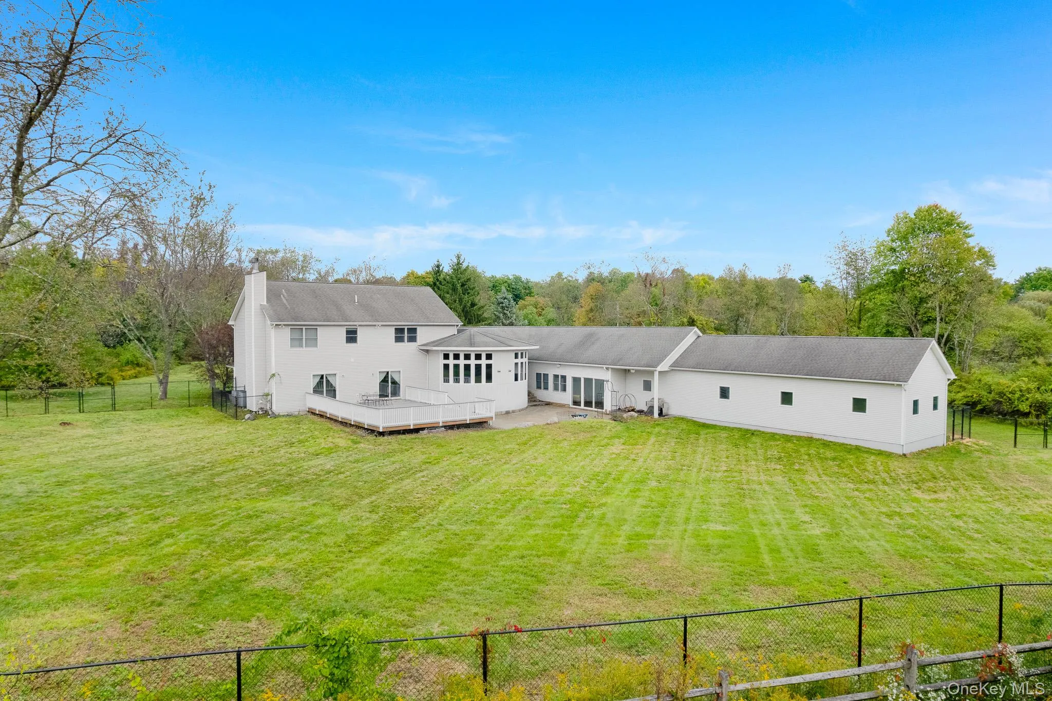 Expansive green lawn with a white residential structure, a white deck, a sunroom with large windows, and a long white auxiliary building. Expansive green lawn with a white residential structure, a white deck, a sunroom with large windows, and a long white auxiliary building.