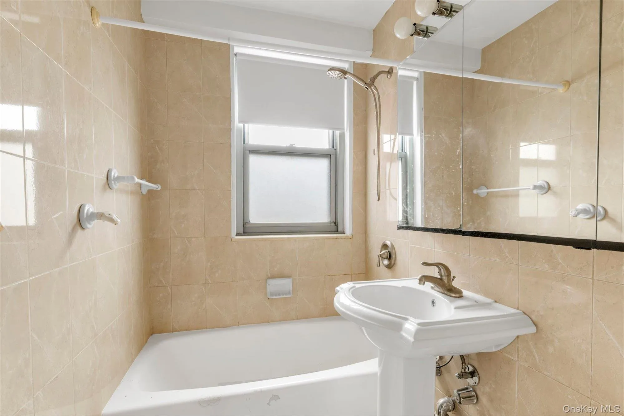 Bathroom featuring a pedestal sink, built-in bathtub with shower head, and tiled walls Bathroom featuring a pedestal sink, built-in bathtub with shower head, and tiled walls
