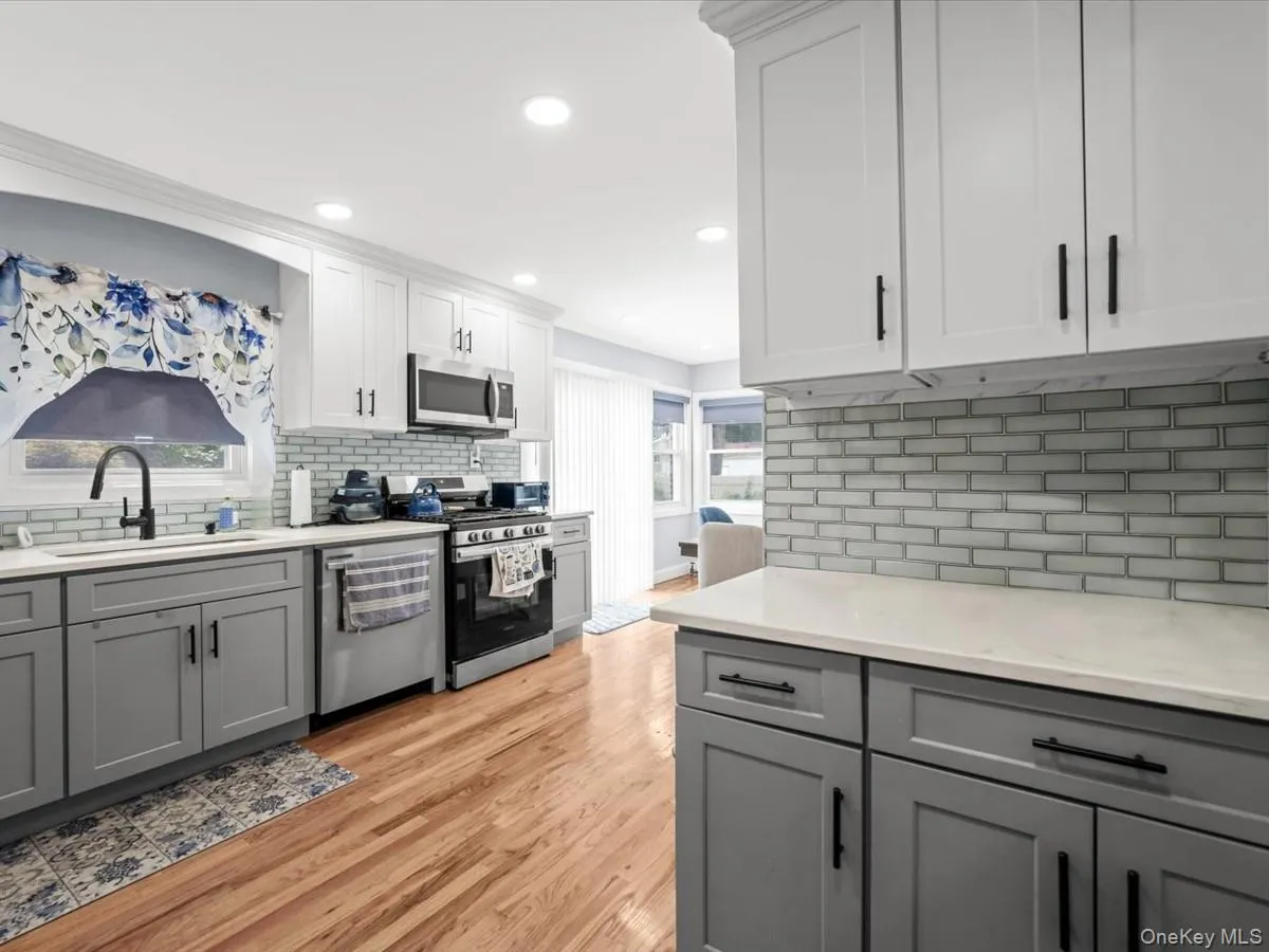 Two-tone cabinetry with white upper and gray lower cabinets, light-toned countertops, subway tile backsplash, stainless steel appliances, and wood-finish flooring Two-tone cabinetry with white upper and gray lower cabinets, light-toned countertops, subway tile backsplash, stainless steel appliances, and wood-finish flooring