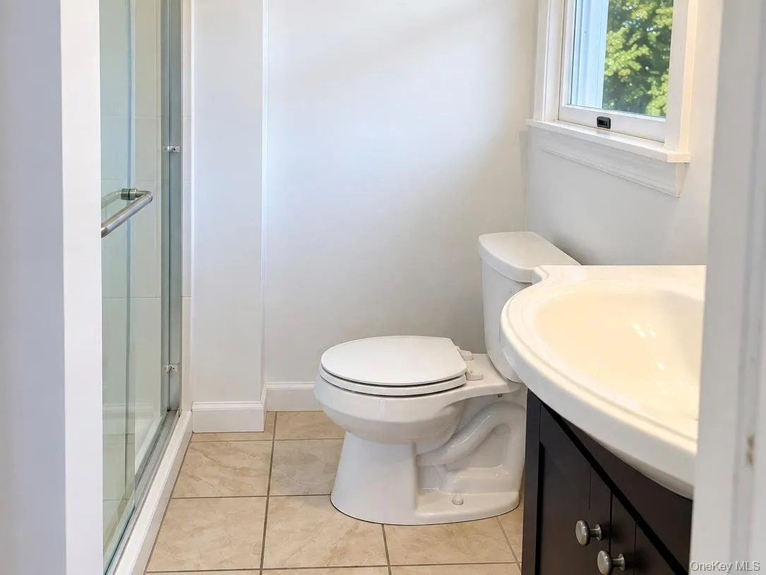 Glass-enclosed shower, white ceramic toilet, integrated sink with dark wood-finish vanity, and tiled flooring Glass-enclosed shower, white ceramic toilet, integrated sink with dark wood-finish vanity, and tiled flooring