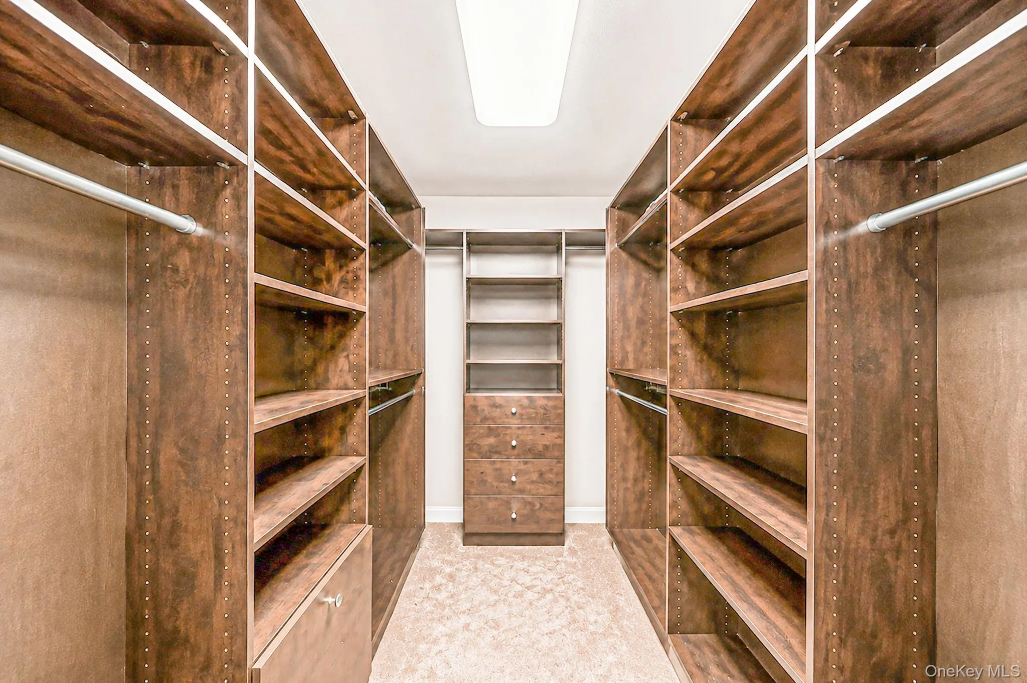 Luxurious walk-in closet featuring rich wood-finish cabinetry with multiple shelving sections, integrated LED lighting throughout, a central dresser with drawer storage Luxurious walk-in closet featuring rich wood-finish cabinetry with multiple shelving sections, integrated LED lighting throughout, a central dresser with drawer storage
