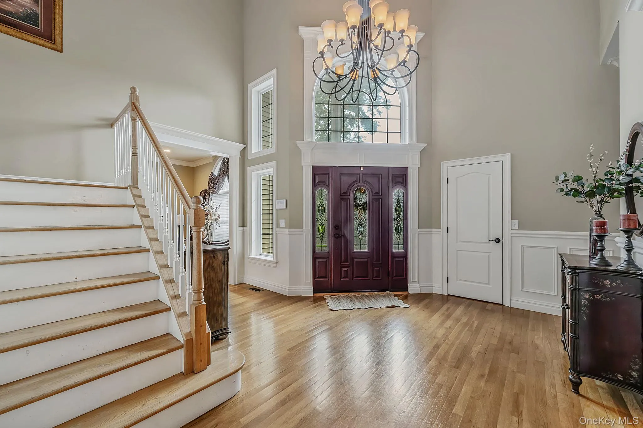 Grand foyer featuring a dual-pane entry door with decorative glass, hardwood flooring, a prominent staircase with wood treads and white risers, an ornate chandelier, and extensive wainscoting Grand foyer featuring a dual-pane entry door with decorative glass, hardwood flooring, a prominent staircase with wood treads and white risers, an ornate chandelier, and extensive wainscoting