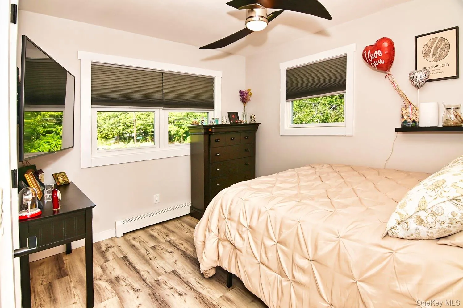 Bright secondary bedroom featuring multiple windows with cellular shades, wood-finish flooring, ceiling fan with integrated lighting, dark wood dresser, and built-in wall shelving