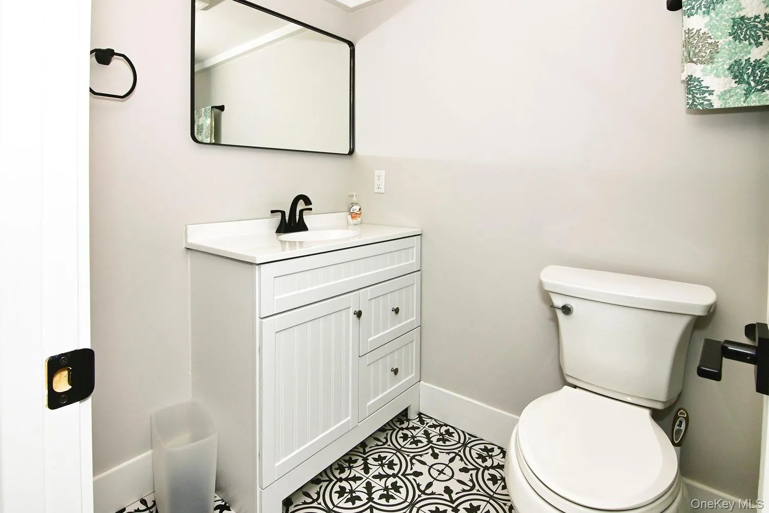 Renovated bathroom featuring a white vanity with multiple drawers and cabinet storage, white porcelain sink, matte black hardware and faucet, ceramic tile flooring with geometric patterning, and a white toilet with elongated bowl