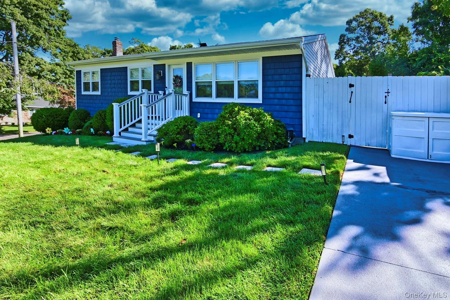 Blue vinyl siding, white trim windows, front entry porch with white railings, mature front landscaping, and a paved driveway