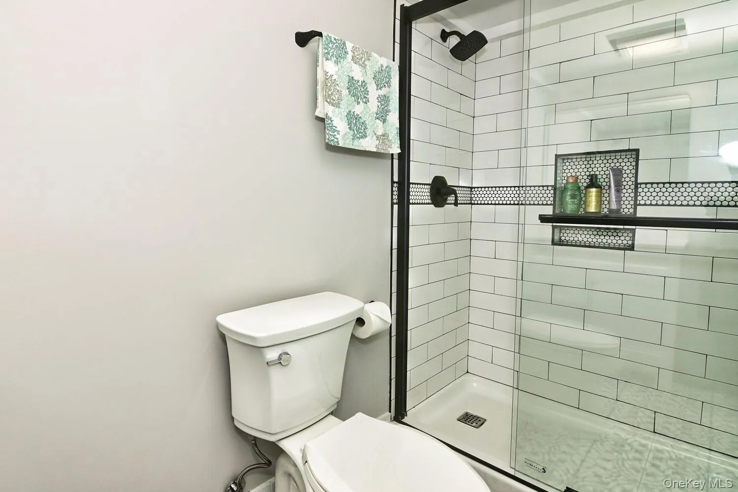 Modern bathroom featuring subway tile walls with contrasting mosaic accent band, frameless glass shower enclosure with brushed black hardware, white porcelain toilet with chrome fixtures, and integrated black metal shelving for storage and display