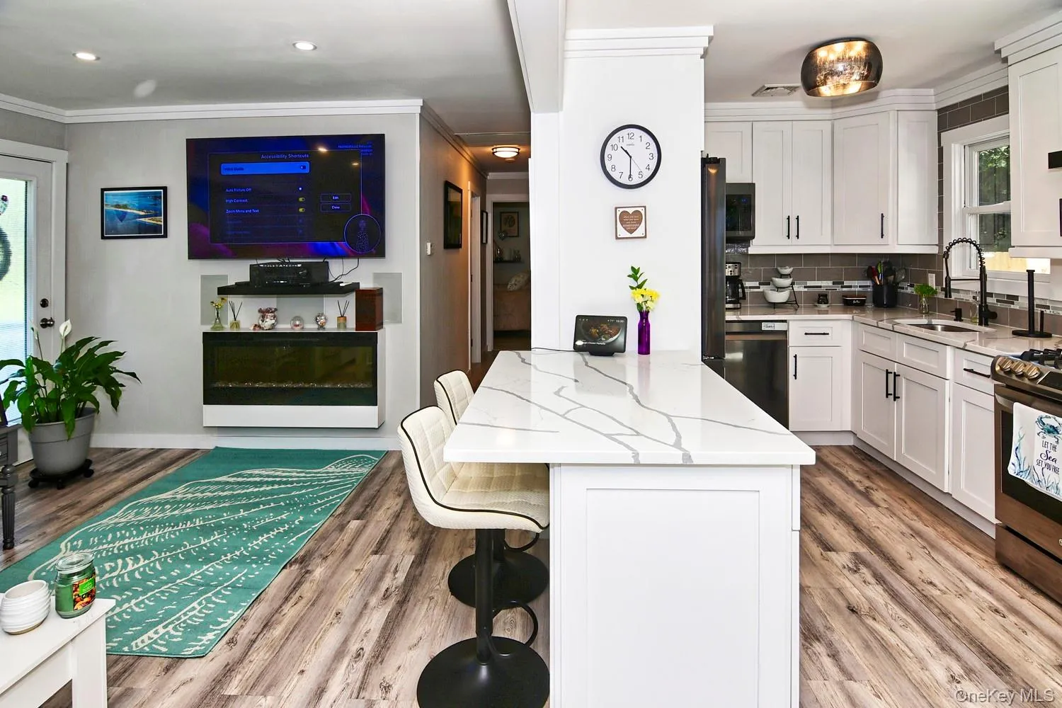 Open-concept kitchen and living area featuring white cabinetry with hardware accents, stainless steel appliances, wood-finish flooring throughout, a marble-look island with seating, and integrated entertainment with wall-mounted display and built-in shelving