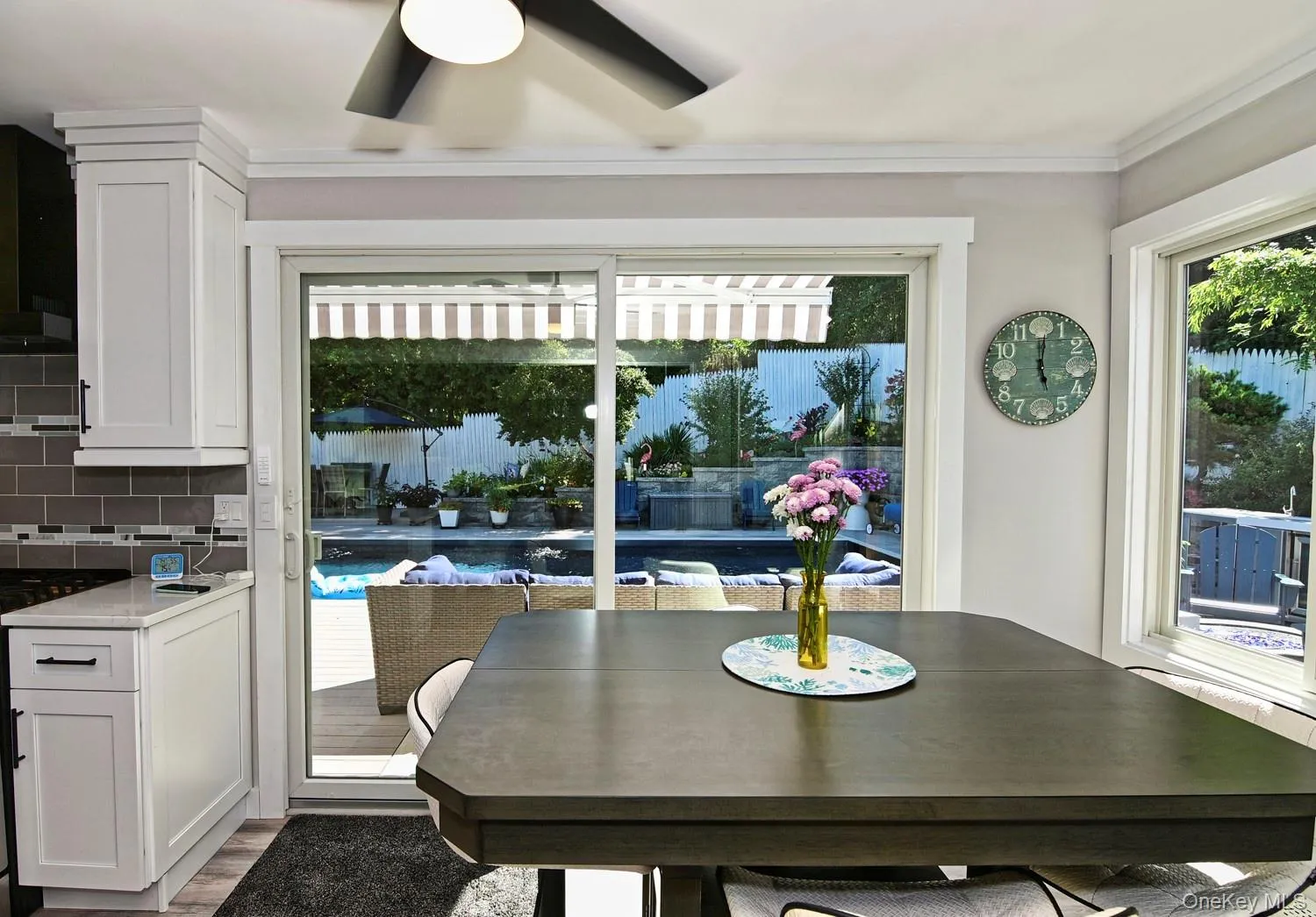 Modern kitchen and dining area featuring sliding glass doors with integrated blinds opening to waterfront views, dark wood-finish dining table with seating for multiple guests, white cabinetry with stainless steel appliances, subway tile backsplash, and abundant natural light from expansive glazing
