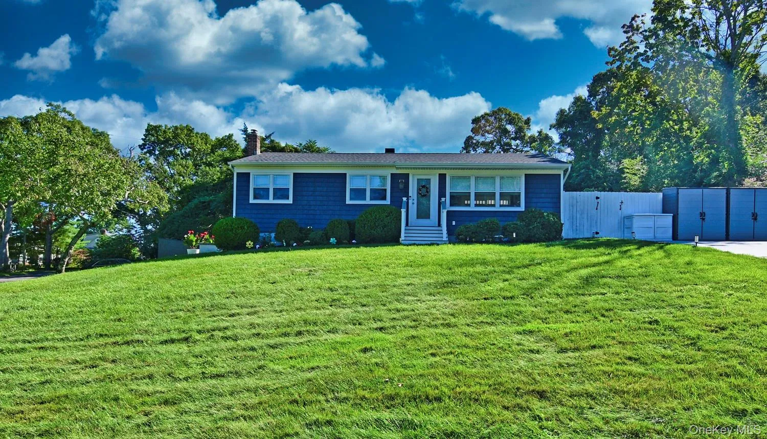 Blue vinyl siding, white trim windows, front entry porch with white railings, mature front landscaping, and a paved driveway