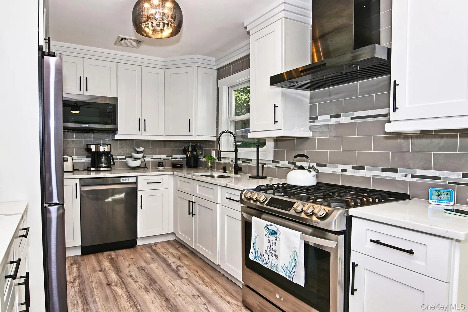 Modern kitchen featuring white shaker cabinetry, stainless steel appliances including a dual-fuel range with five burners, quartz countertops, subway tile backsplash, black range hood, and wood-finish flooring throughout