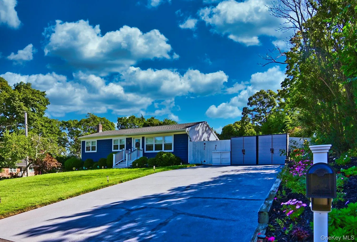 Blue vinyl siding, white trim windows, front entry porch with white railings, mature front landscaping, and a paved driveway