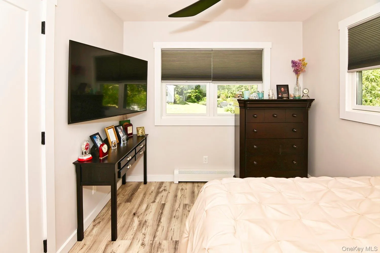 Bright secondary bedroom featuring wood-finish flooring, a wall-mounted flat-screen television, large windows with cellular shading allowing natural light, dark wood dresser with multiple drawers, and a ceiling fan