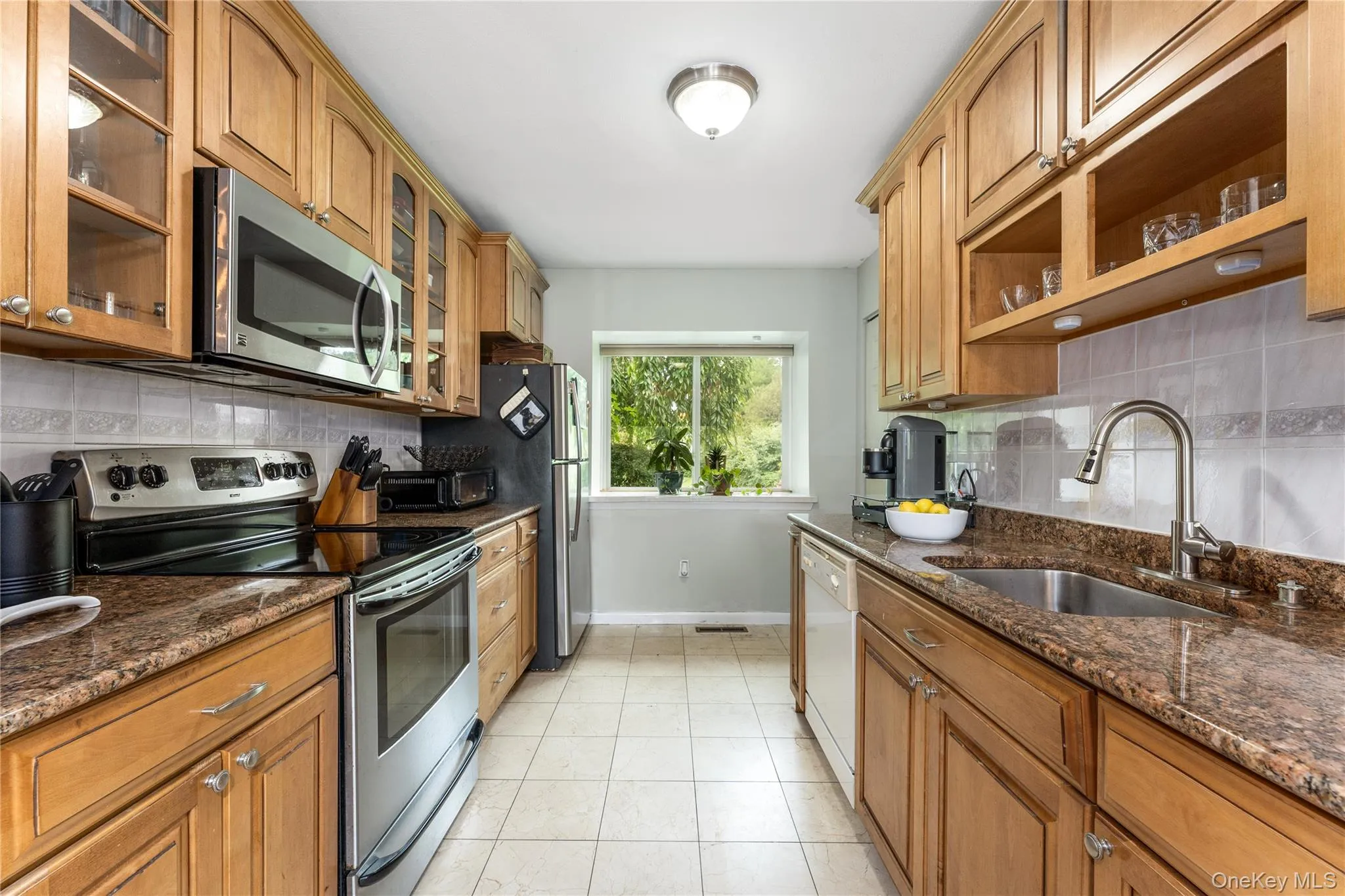 Galley kitchen featuring light wood cabinetry, stainless steel appliances, speckled countertops, tile flooring, and a tiled backsplash Galley kitchen featuring light wood cabinetry, stainless steel appliances, speckled countertops, tile flooring, and a tiled backsplash