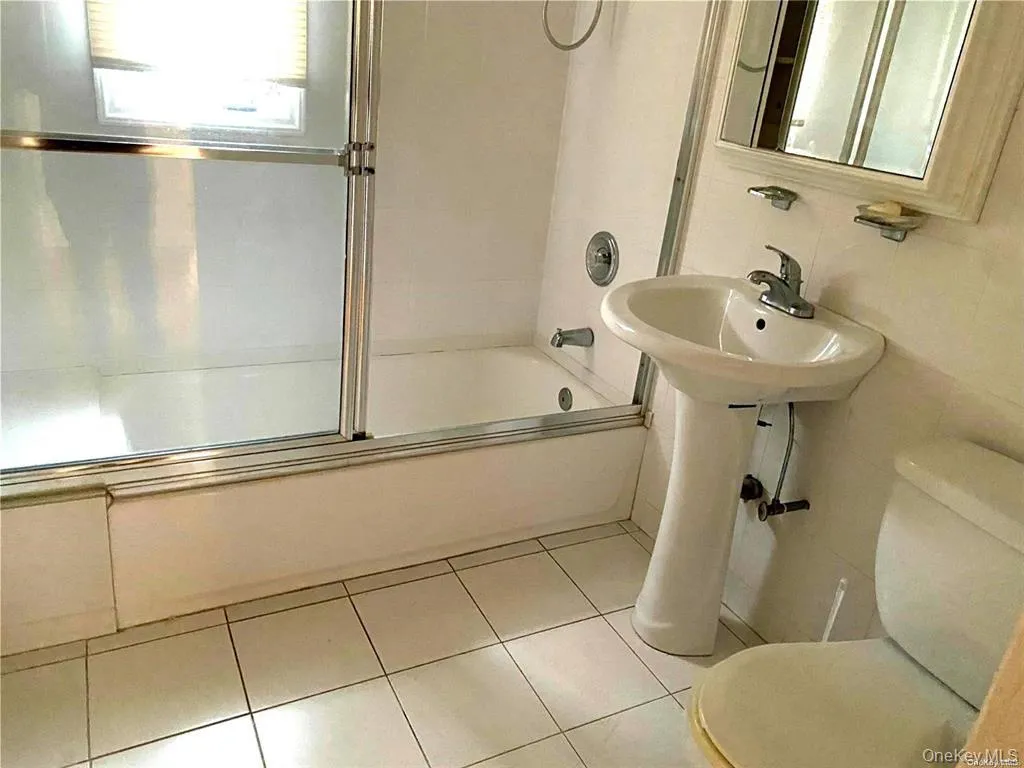 Pedestal sink with chrome faucet, white tiled flooring, bathtub with sliding glass enclosure, white wall tiling, and mirrored vanity cabinet Pedestal sink with chrome faucet, white tiled flooring, bathtub with sliding glass enclosure, white wall tiling, and mirrored vanity cabinet