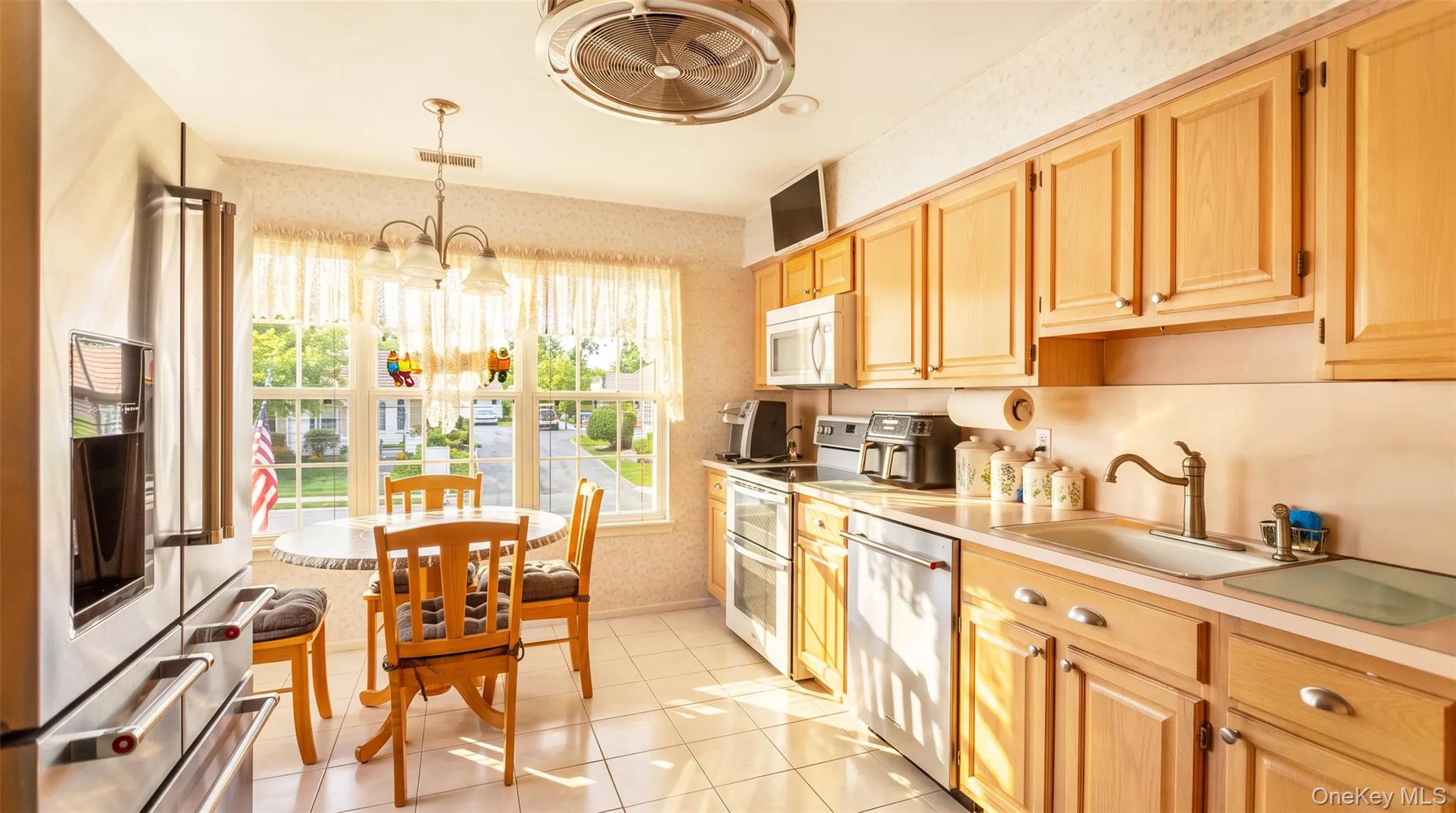 Kitchen featuring light wood cabinetry, stainless steel appliances, tile flooring, and a dining area with a multi-pane window Kitchen featuring light wood cabinetry, stainless steel appliances, tile flooring, and a dining area with a multi-pane window