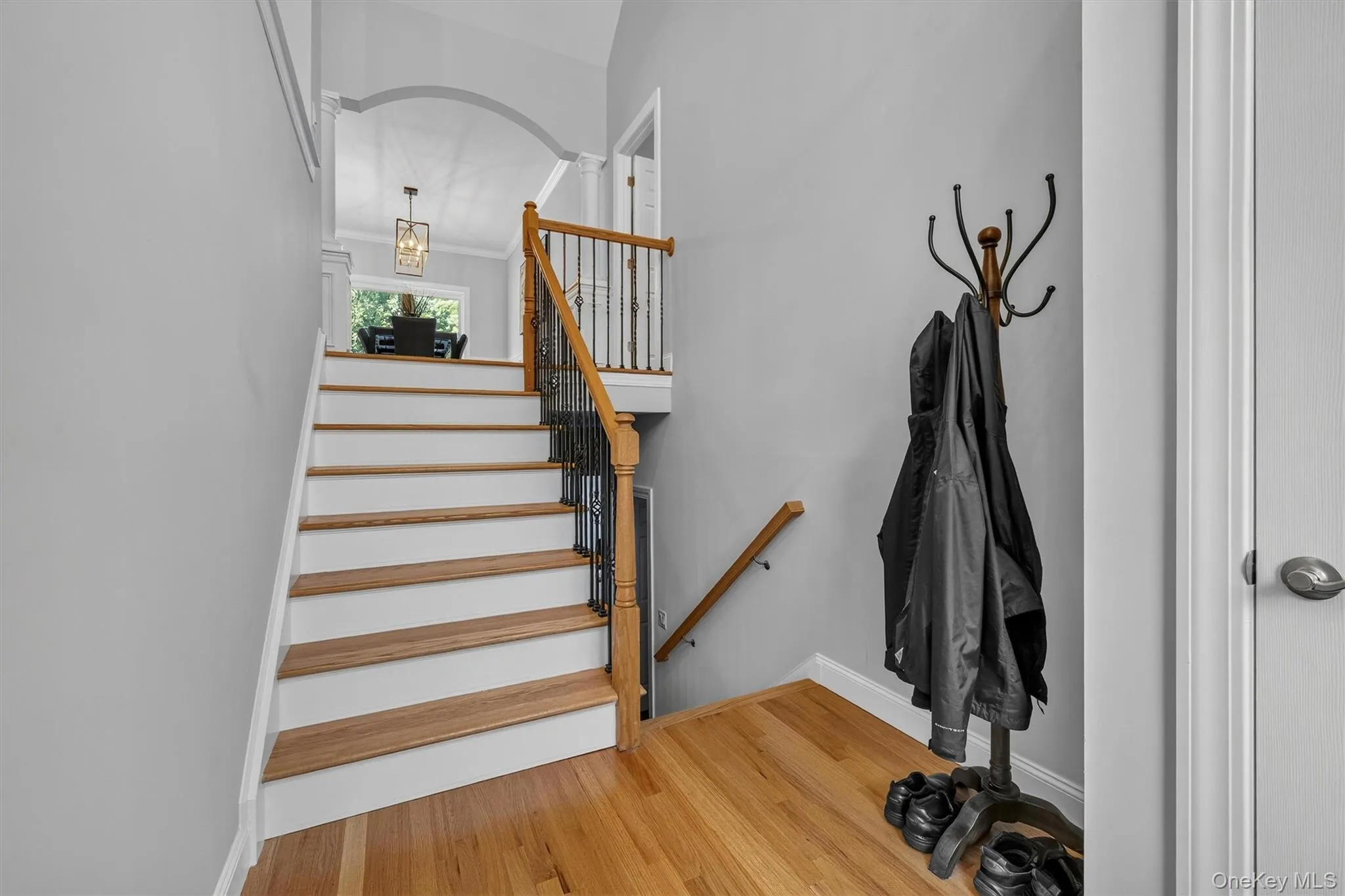 Welcoming entry foyer featuring a striking arched doorway, wide wooden staircase with wooden handrail and metal balusters, warm wood-finish flooring, and a wall-mounted coat rack with hooks for convenient storage Welcoming entry foyer featuring a striking arched doorway, wide wooden staircase with wooden handrail and metal balusters, warm wood-finish flooring, and a wall-mounted coat rack with hooks for convenient storage