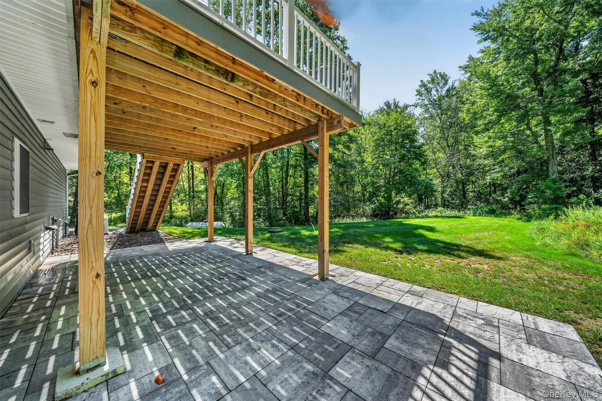 Expansive paver patio with a covered outdoor area, wooden deck above, and a lush green lawn Expansive paver patio with a covered outdoor area, wooden deck above, and a lush green lawn