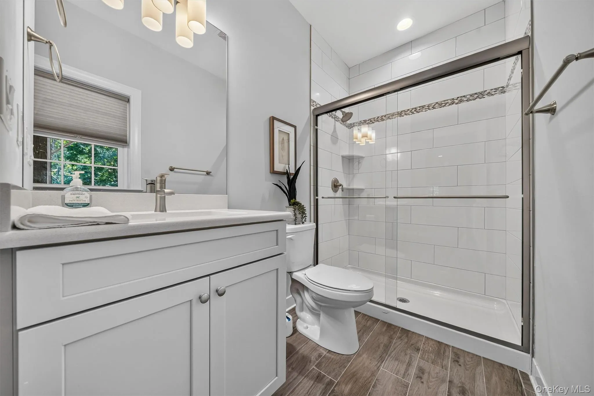 Wood-finish flooring, white vanity with integrated sink, rectangular mirror, glass-enclosed shower with subway tile, and a decorative tile accent band Wood-finish flooring, white vanity with integrated sink, rectangular mirror, glass-enclosed shower with subway tile, and a decorative tile accent band