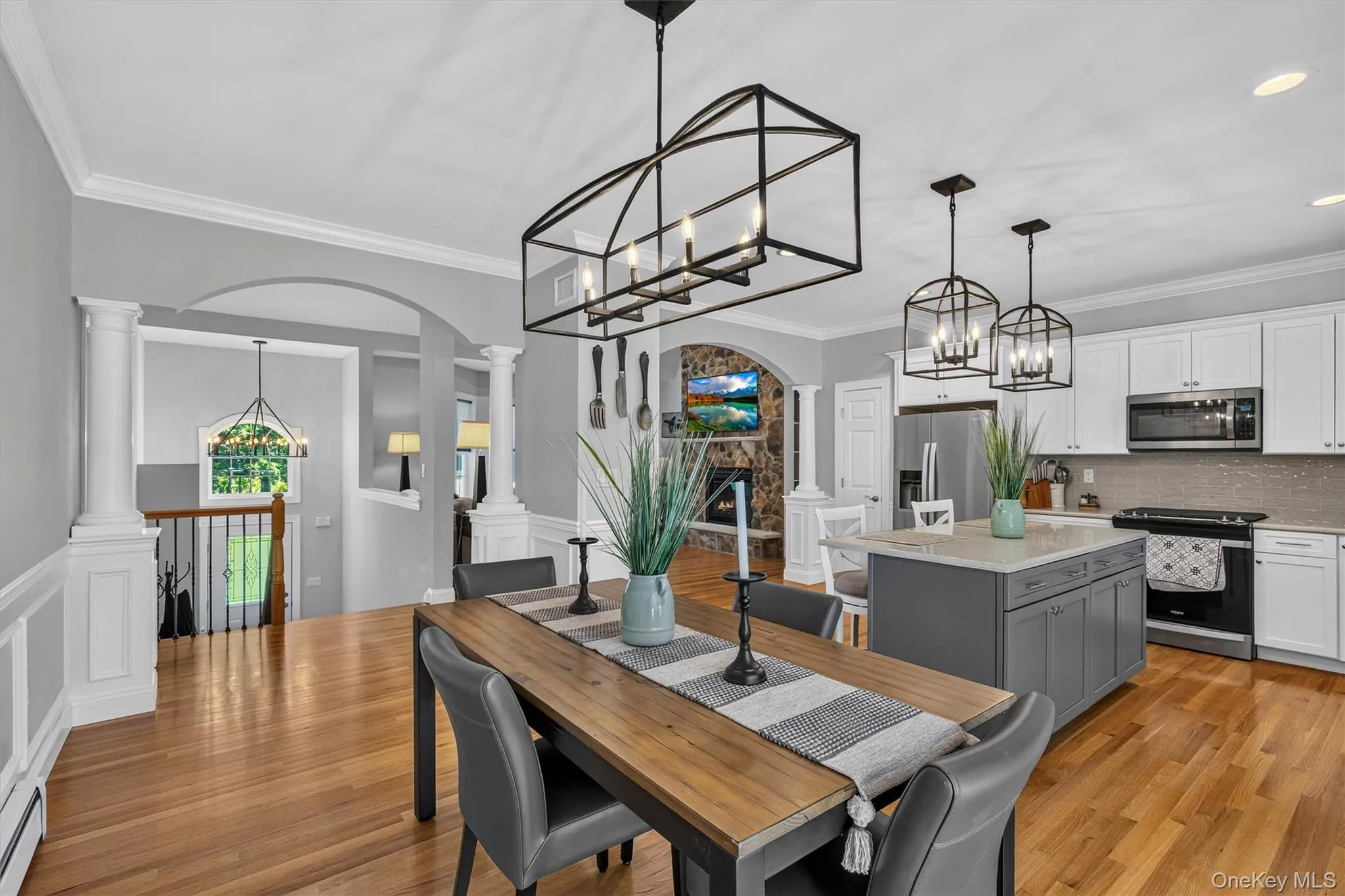 Open-concept dining and kitchen area featuring hardwood flooring, custom cabinetry with gray-painted island, stainless steel appliances, and pendant lighting Open-concept dining and kitchen area featuring hardwood flooring, custom cabinetry with gray-painted island, stainless steel appliances, and pendant lighting