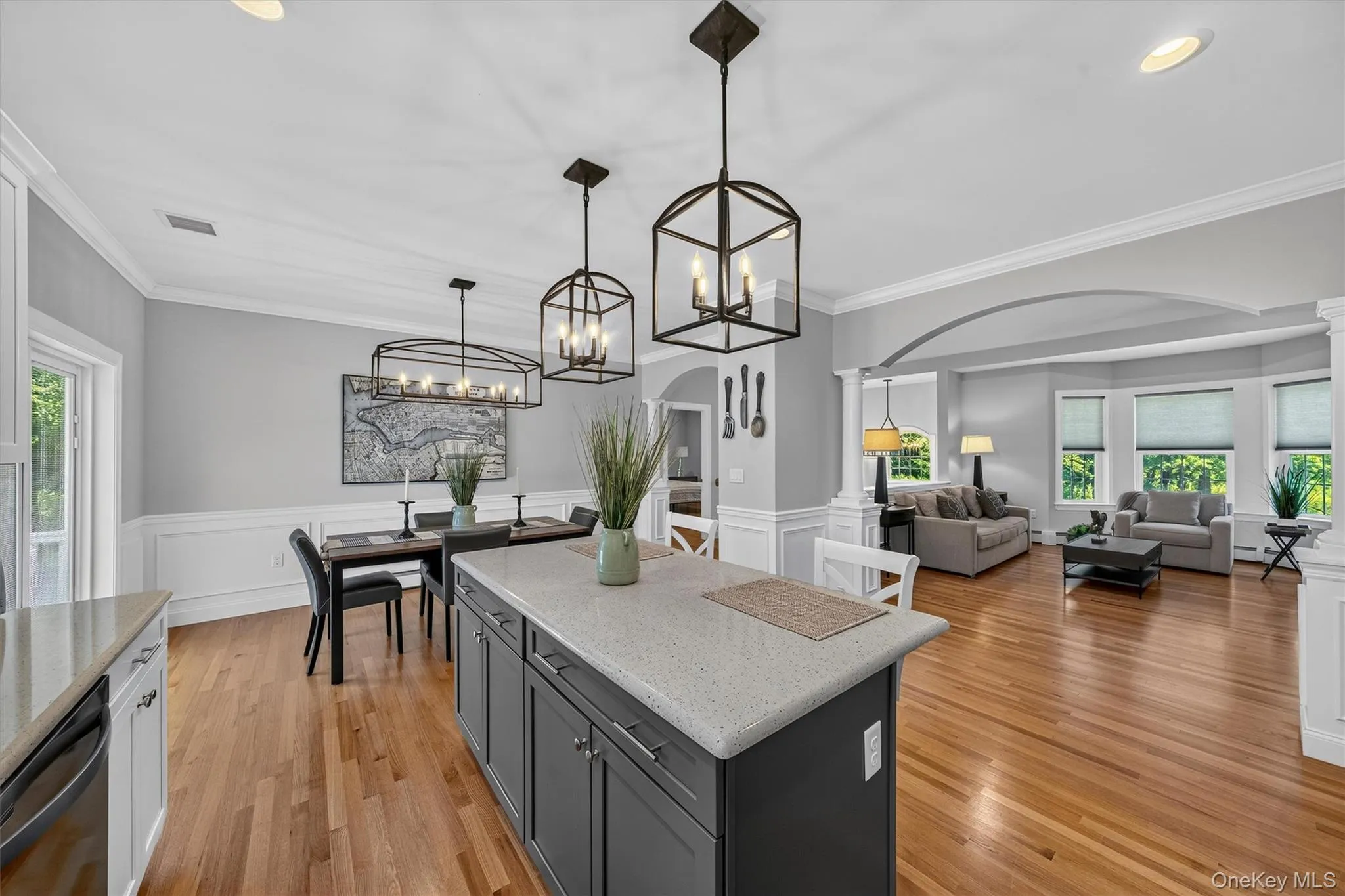 Open-concept kitchen and dining area featuring a large island with quartz countertop and dark cabinetry, pendant lighting, hardwood flooring, and seamless sightlines into the adjoining living spaces accented by wainscoting and architectural archways Open-concept kitchen and dining area featuring a large island with quartz countertop and dark cabinetry, pendant lighting, hardwood flooring, and seamless sightlines into the adjoining living spaces accented by wainscoting and architectural archways