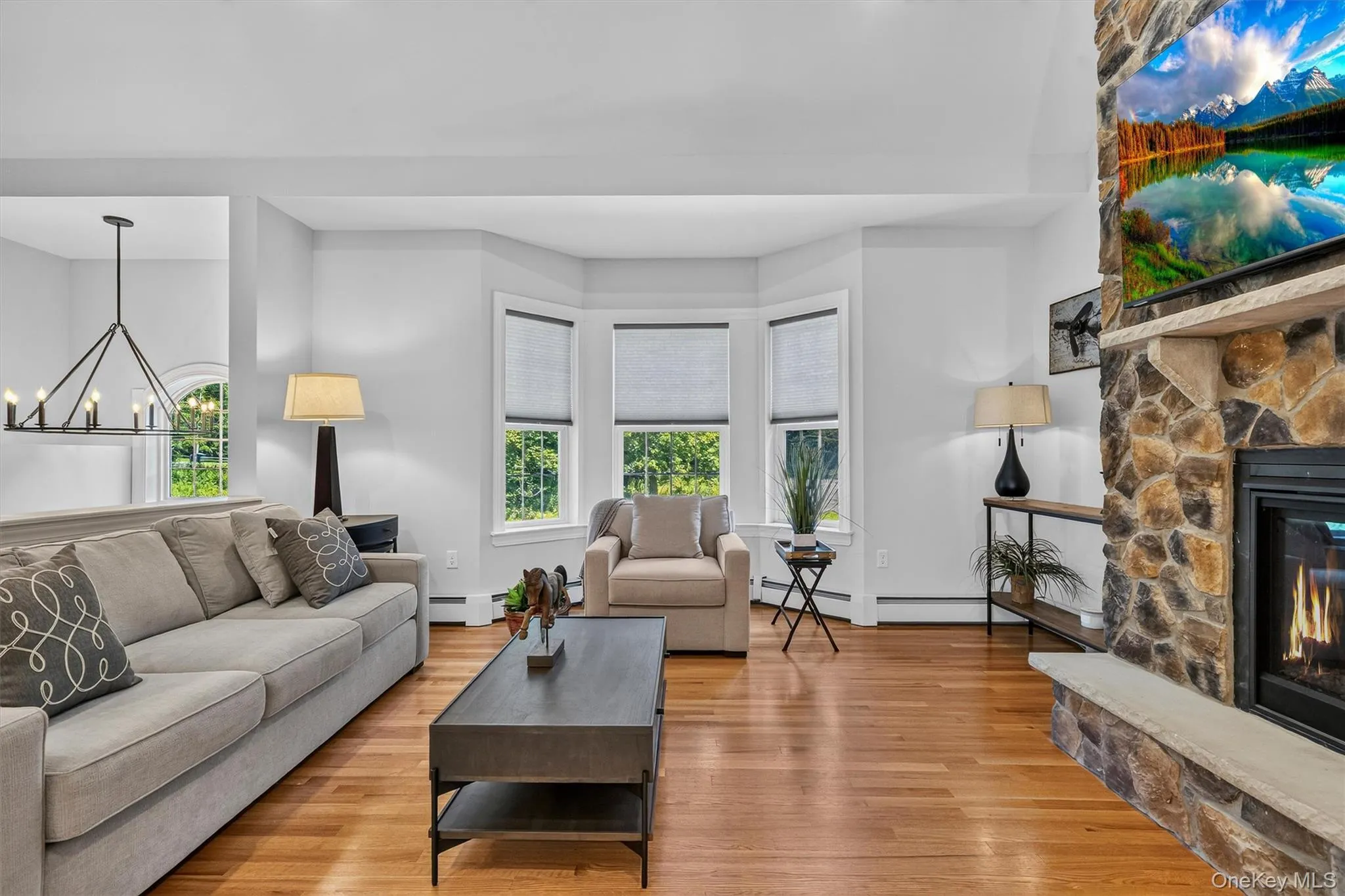 Bright, light-filled living room featuring bay windows with multiple panes, hardwood flooring, a stone fireplace with gas insert, a dark wood coffee table with lower shelf storage, pendant lighting, and neutral wall finishes throughout Bright, light-filled living room featuring bay windows with multiple panes, hardwood flooring, a stone fireplace with gas insert, a dark wood coffee table with lower shelf storage, pendant lighting, and neutral wall finishes throughout