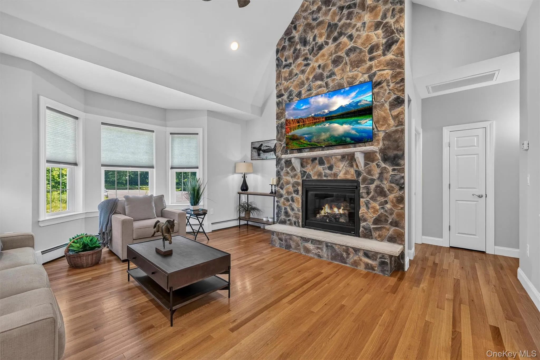 Bright living room featuring hardwood flooring, a striking stone fireplace with gas insert and mounted television, bay windows with multiple panes, recessed ceiling lighting, and white trim throughout Bright living room featuring hardwood flooring, a striking stone fireplace with gas insert and mounted television, bay windows with multiple panes, recessed ceiling lighting, and white trim throughout