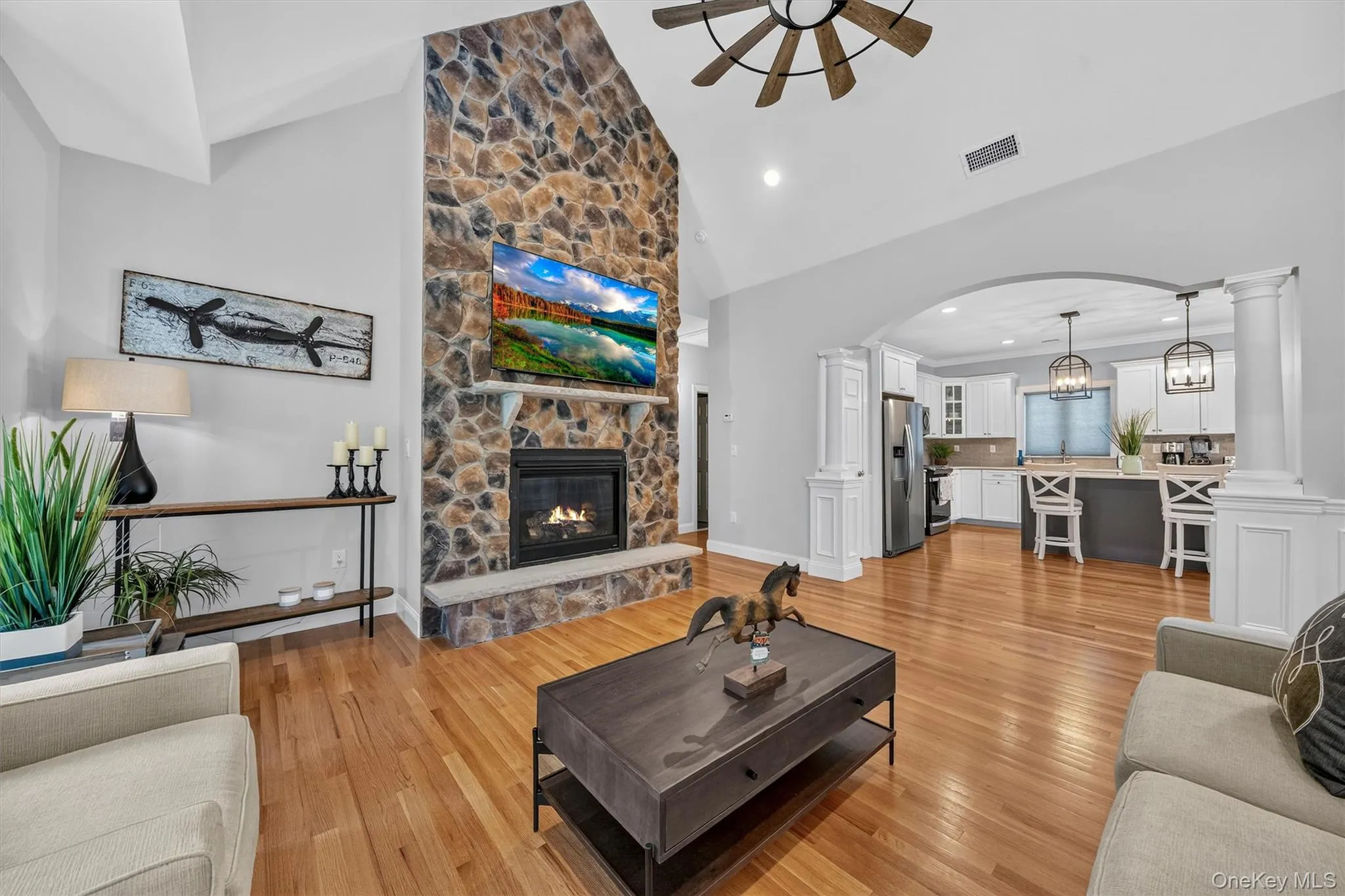 Vaulted ceiling with a ceiling fan, stone-surround fireplace with a mantel, wood-finish flooring, and an arched entryway Vaulted ceiling with a ceiling fan, stone-surround fireplace with a mantel, wood-finish flooring, and an arched entryway