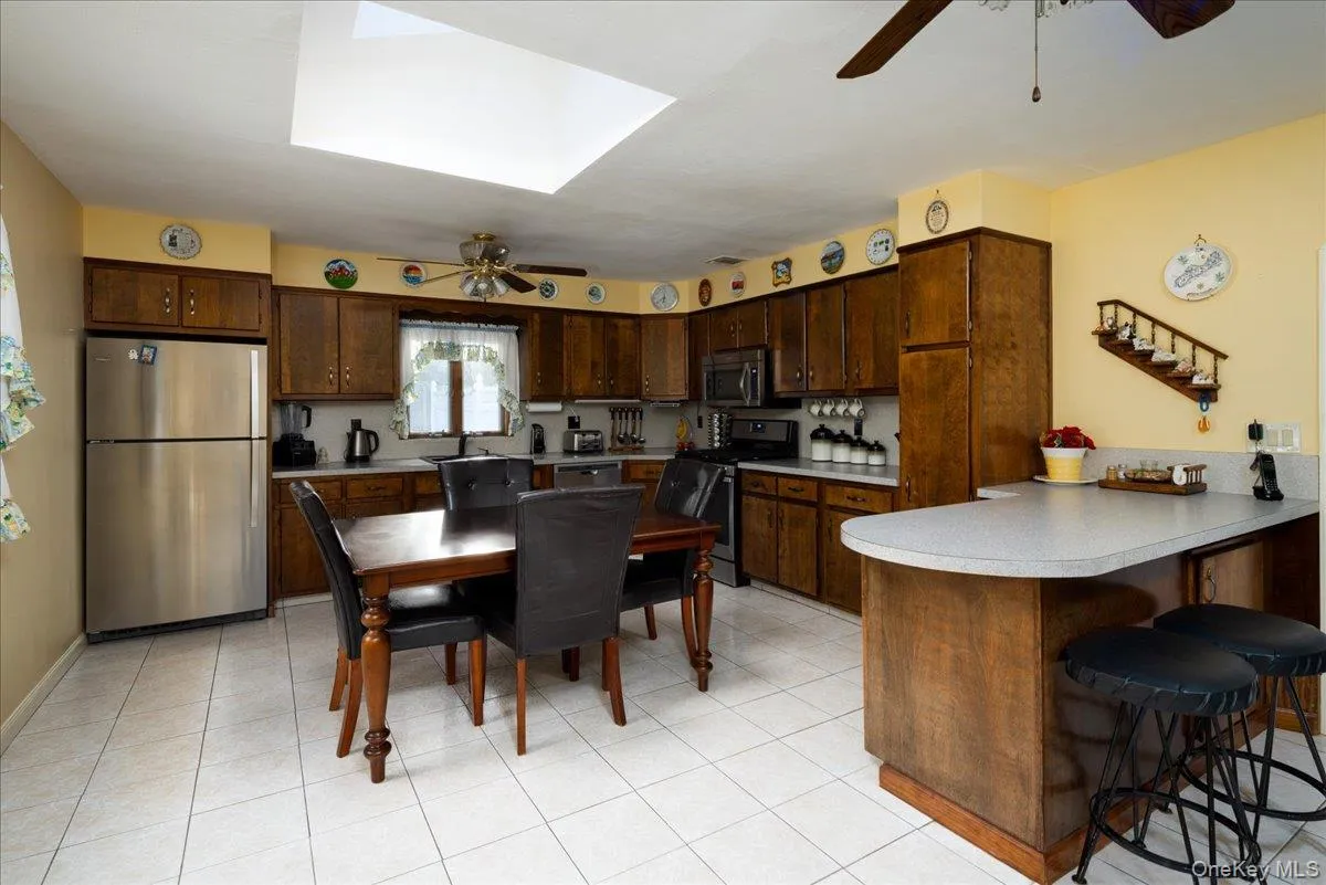 Kitchen featuring wood cabinetry, stainless steel appliances, white tile flooring, a skylight, and a built-in breakfast bar Kitchen featuring wood cabinetry, stainless steel appliances, white tile flooring, a skylight, and a built-in breakfast bar
