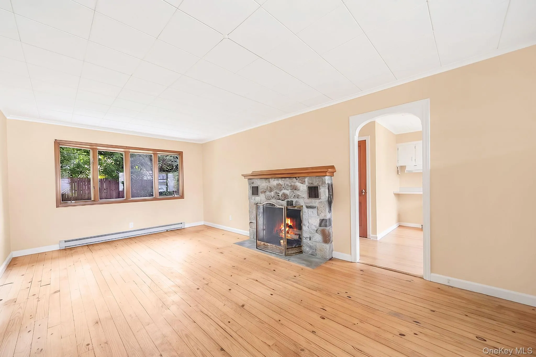Stone fireplace with wood mantel, light wood-finish flooring, large window with wood trim, archway entry, and white ceiling panels Stone fireplace with wood mantel, light wood-finish flooring, large window with wood trim, archway entry, and white ceiling panels