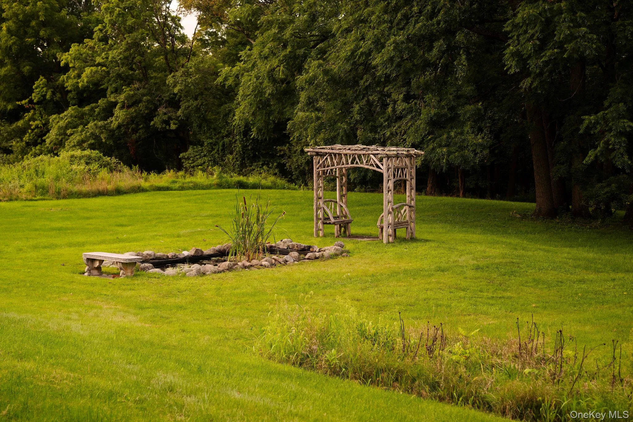 Expansive grassy lawn with a rock-lined pond, stone bench, and an ornate wooden arbor featuring two swings Expansive grassy lawn with a rock-lined pond, stone bench, and an ornate wooden arbor featuring two swings