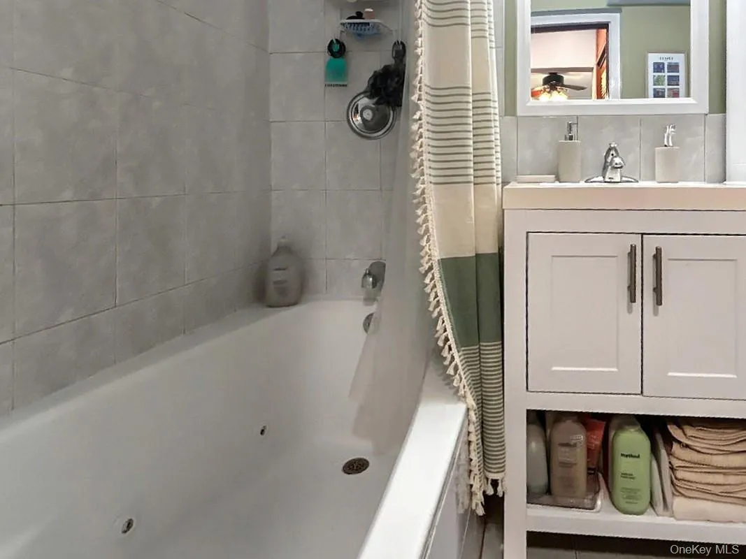 Bathtub with light-colored tile surround, white vanity with dual doors, open shelving, and a wall-mounted mirror plus additional storage above shower Bathtub with light-colored tile surround, white vanity with dual doors, open shelving, and a wall-mounted mirror plus additional storage above shower
