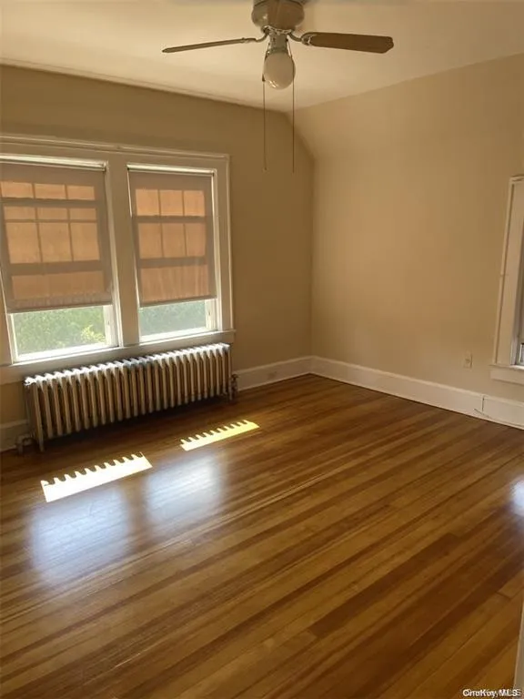 Wood-finish flooring, dual windows with roller shades, ceiling fan with integrated lighting, and a wall-mounted radiator Wood-finish flooring, dual windows with roller shades, ceiling fan with integrated lighting, and a wall-mounted radiator