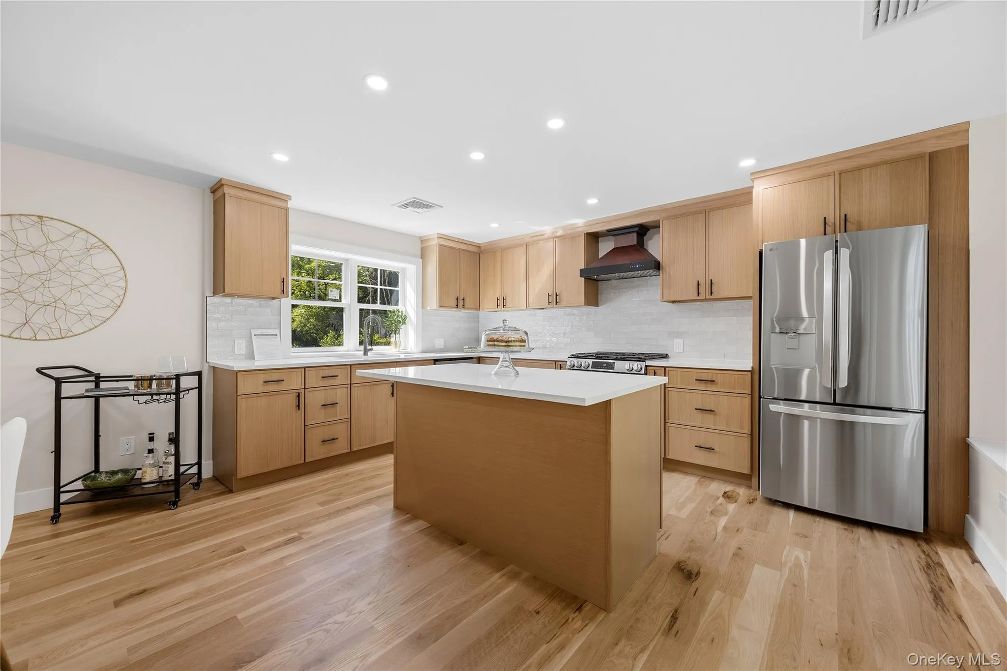 Spacious kitchen featuring light wood cabinetry, a central island with a white countertop, stainless steel appliances, white subway tile backsplash, and wood-finish flooring Spacious kitchen featuring light wood cabinetry, a central island with a white countertop, stainless steel appliances, white subway tile backsplash, and wood-finish flooring