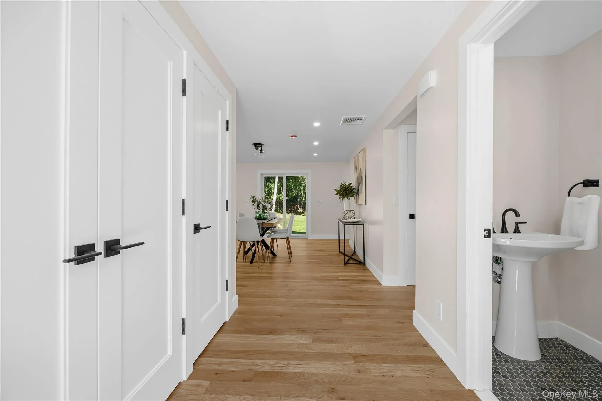 Light wood-finish flooring, recessed lighting, white interior doors with black hardware, white pedestal sink with black faucet, and dark circular tile flooring Light wood-finish flooring, recessed lighting, white interior doors with black hardware, white pedestal sink with black faucet, and dark circular tile flooring